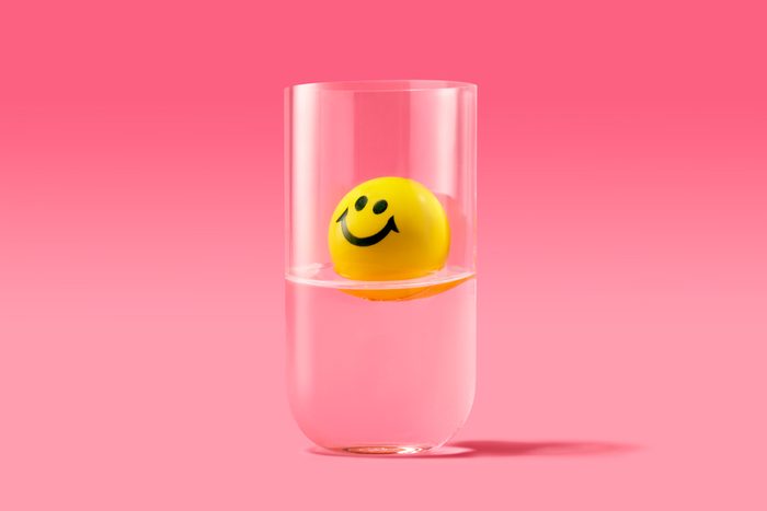 yellow smiley face looking upwards optimistically while floating in a half full glass of water; pink background