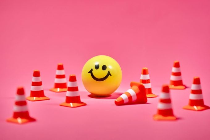 a yellow smiley face ball surrounded by traffic boundary cones on pink background; one cone has fallen over onto its side