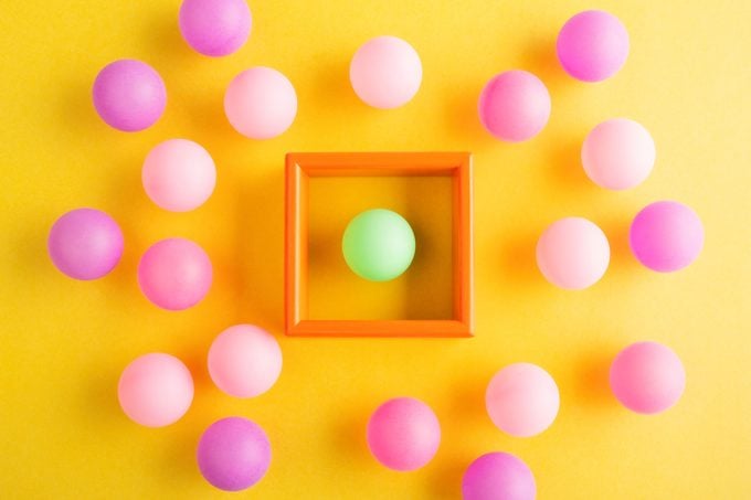One little green ball within a wooden box, many pink balls outside the box on yellow surface, view from directly above
