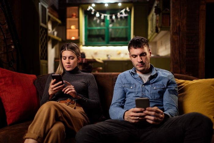 a young couple sit on the couch and look at their phones ignoring each other