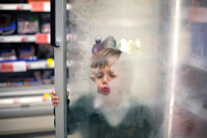 A little boy is keeping himself busy with melting with his breath the ice on the glass door of a refrigerator in a supermarket during the shopping trip.
