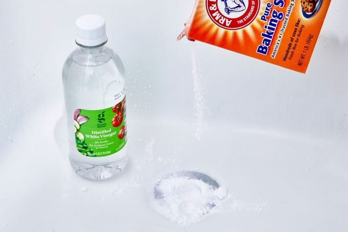 pouring baking soda into a bathtub drain, bottle of white vinegar nearby