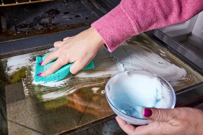 hand using baking soda and dishsoap paste to scrub the interior of a glass oven door with a sponge
