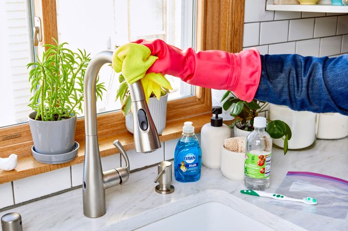 anonymous hand cleaning kitchen sink faucet; cleaning supplies are seen on the counter nearby