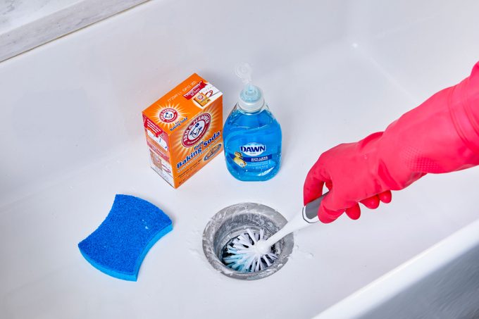 hand using scrubbing brush to clean kitchen sink garbage disposal; cleaning supplies sit nearby