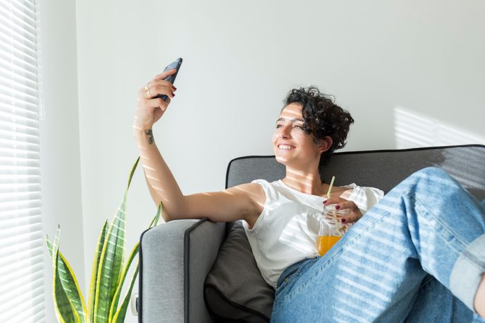 Smiling young woman sitting on couch with a soft drink taking a selfie