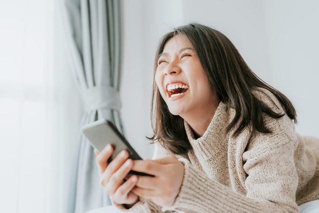 Happy woman laughing while lying on the bed with smartphone