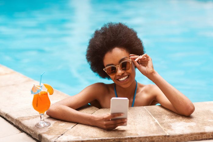 Beautiful black girl with tropical cocktail browsing internet on smartphone at swimming pool