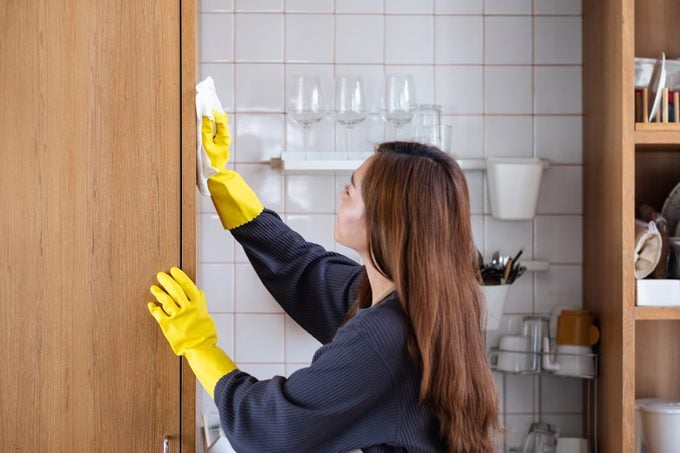 A woman wearing protective glove, cleaning wooden cupboard in the kitchen at home