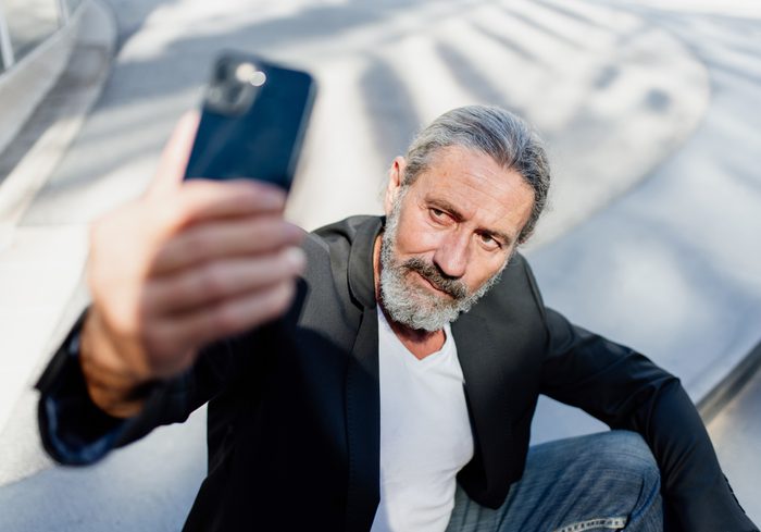 Front view of a middle-aged businessman with ponytail and blazer takes a selfie with his smartphone in the city