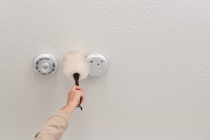 Woman reaching up to dust smoke alarm on ceiling at home