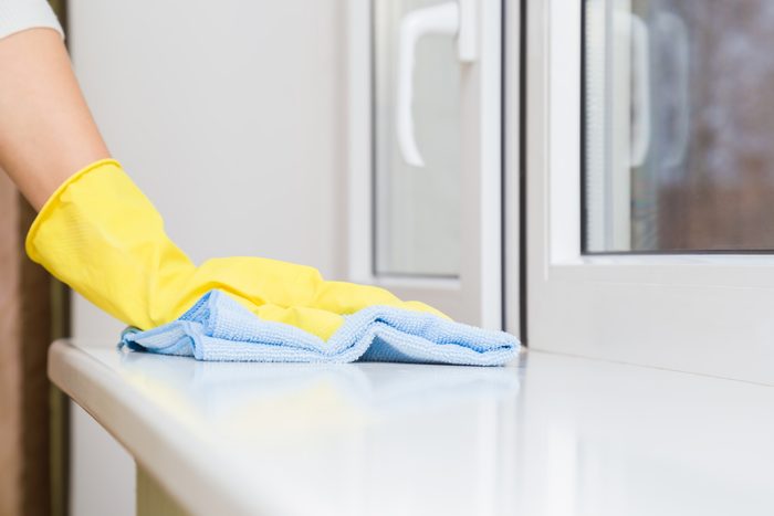 Woman hand in yellow rubber protective glove wiping white plastic window sill from dust with blue dry rag. General or regular cleanup. Cleaning service. Closeup.
