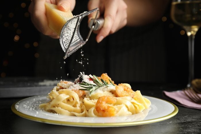 Woman grating cheese onto pasta with shrimps on black table, closeup