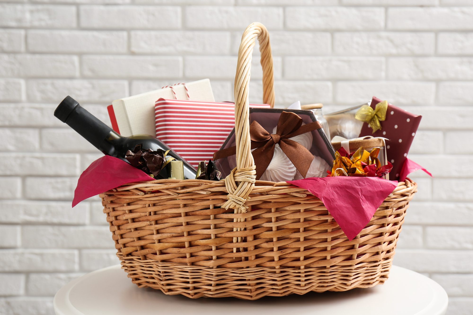 Wicker gift basket with bottle of wine on table near white brick wall