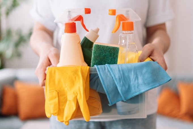 Lady hand holding cleaning products with rags and gloves in a plastic container