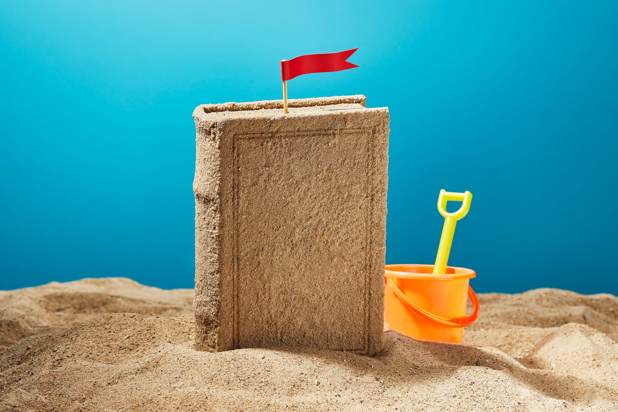 a "sand castle" made in the shape of a book stands up from the sand with a red flag on top. orange pail and yellow shovel is nearby. blue background