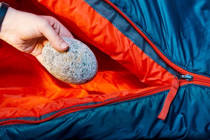 a hand placing a warmed rock into a partially unzipped sleeping bag