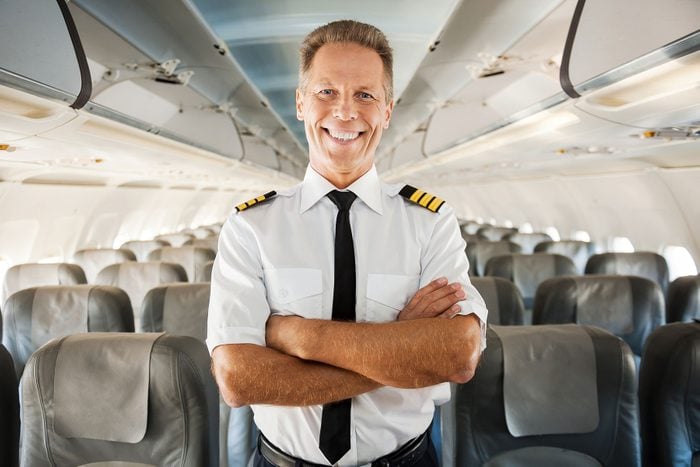 Confident male pilot in uniform keeping arms crossed and smiling while standing inside of the airplane