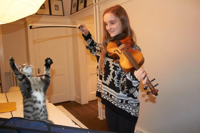 young red headed girl short hair brown eyes and freckles playing violin in the house teasing cat with the bow of violin cat is reaching up to bow trying to grab it above its head girl is smiling