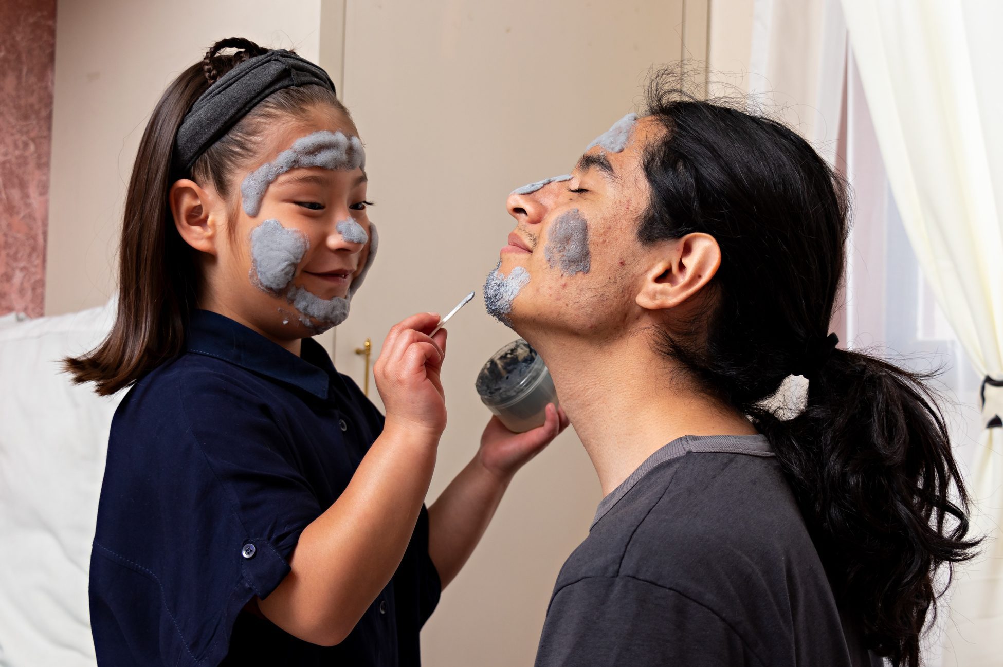 Young man with a girl sitting on a bed talking and laughing, while putting on a face cream, playing in a spa at home