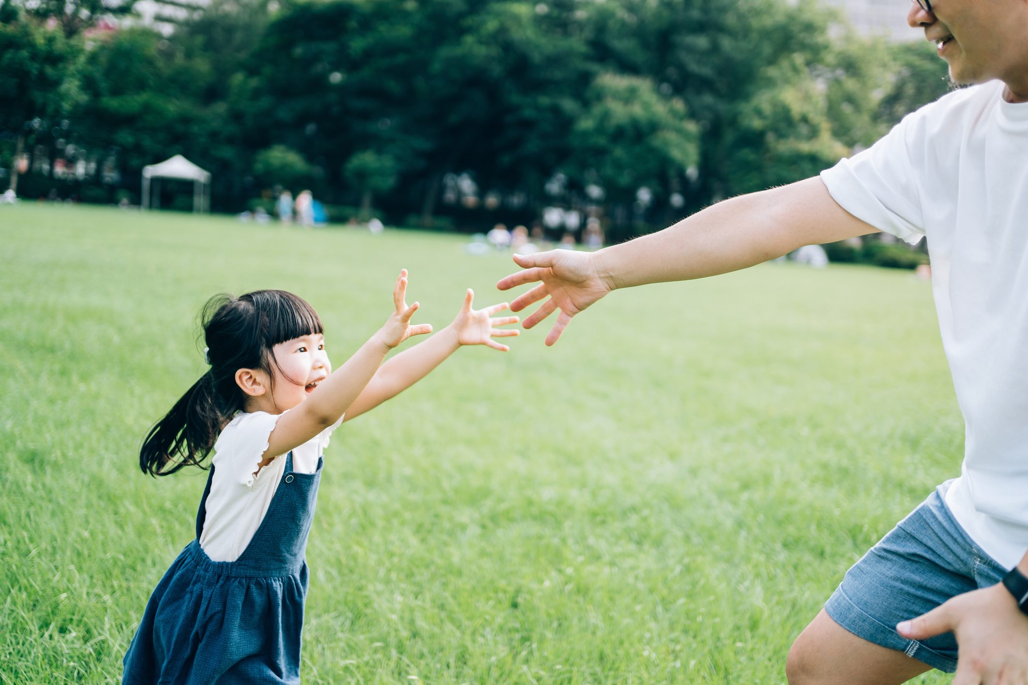 Happy little Asian girl and young Asian father having fun outdoors, running around on lawn. Father is giving his hand to daughter. Enjoying nature and Summer days in urban park. Family love lifestyle. Trust and support concept