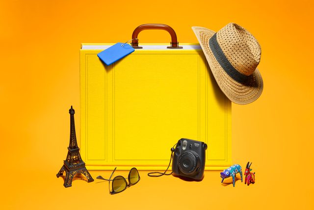 a yellow book stands upright on its spine to appear like a suitcase, with handle and blue luggage tag coming up from the pages, and a hat perched on one corner of the book. souvenirs, sunglasses, and a camera are arranged in front. all on orange background