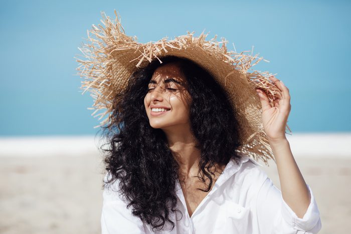 young beautiful mixed race woman with long curly hair sits on the beach in a straw hat with her eyes closed, looks at the sun and smiles