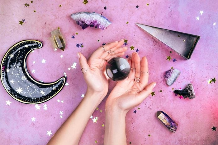 Hands with rings on fingers are holding crystal ball near esoteric set on concrete gray background with many stars sequins. Flat lay style