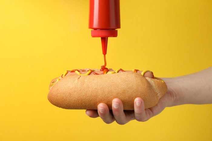Person pouring ketchup on hot dog, on yellow background