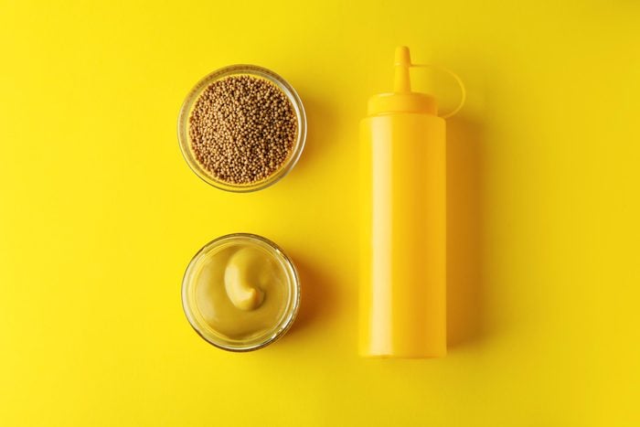 a small bowl of mustard seed, mustard and a plastic bottle of mustard on a yellow background