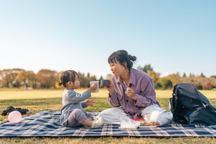 Mother and daughter enjoying having picnic together in public park on warm sunny day