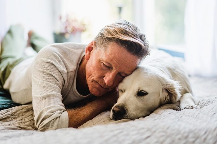 man snuggling with dog on a bed, looking solemn