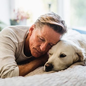 man snuggling with dog on a bed, looking solemn