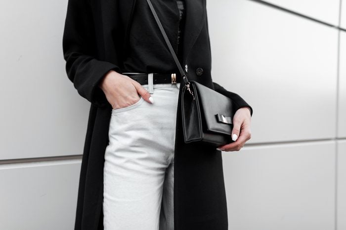 Woman in a trendy T-shirt in a black long fashionable coat in stylish white jeans with a black leather elegant bag. Trendy elegant casual outfit. Details of everyday look. Street fashion. Close-up