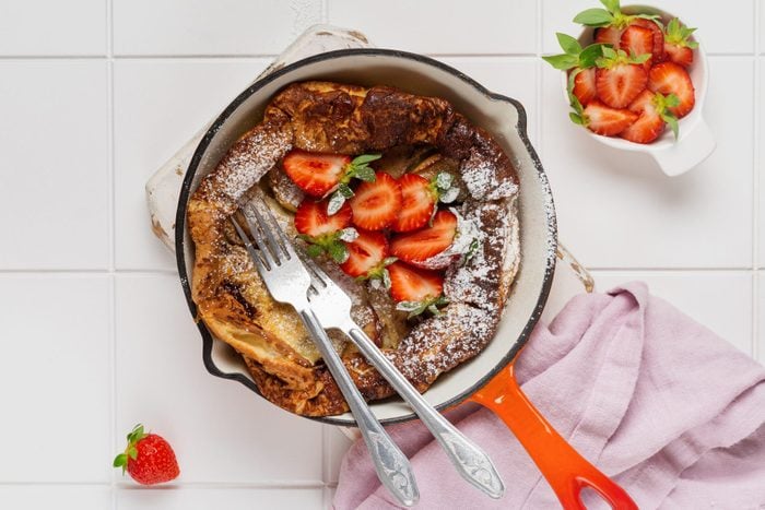 Dutch baby pancake with fresh strawberry berry and sprinkled with icing sugar powder in red pan on white kitchen background. Top view.
