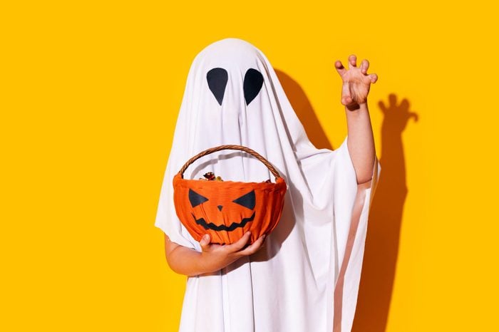 Child in ghost costume holding basket of chocolates in one hand, and raised the other hand up
