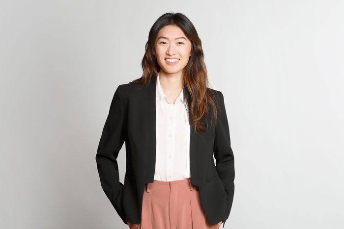 Portrait Of An Asian Millennial Woman With Long Hair, Smiling In Front Of A Grey Backdrop, Wearing A Black Blazer, A White Blouse And Coral Colored Pants.