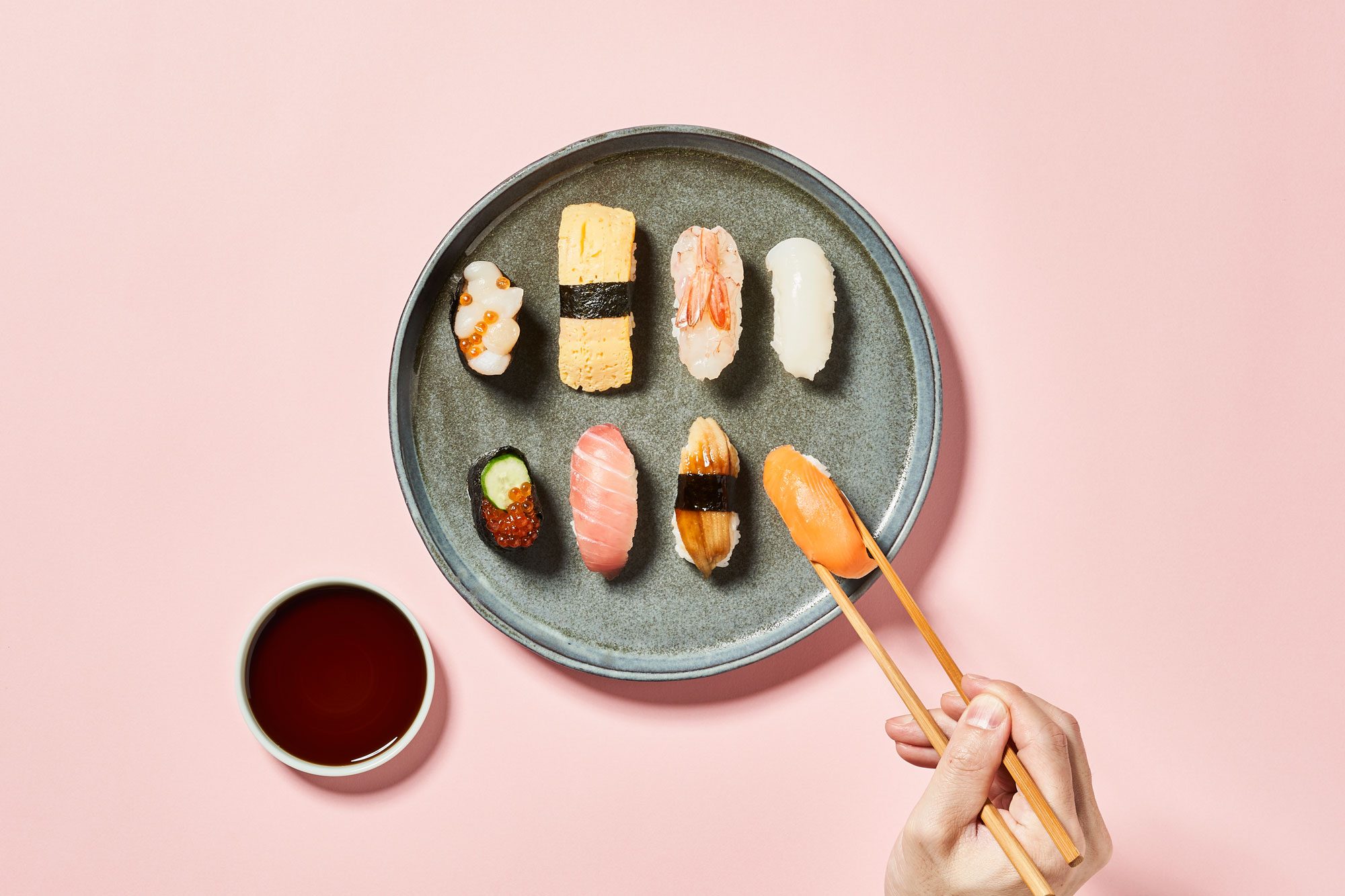 round plate with sushi pieces on pink background. a hand with chopsticks holds a piece of sushi. soy sauce dish nearby