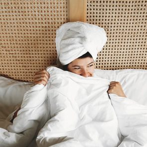 Beautiful happy young woman hiding under white sheets, lying on bed in hotel room or home bedroom. Stylish girl with white towel on hair relaxing on white bed. Space for text