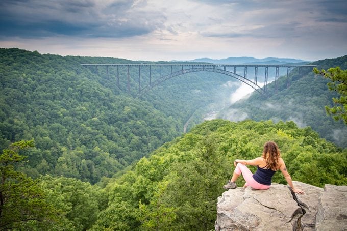 A woman perched on a rocky outcrop looks out over the New River Gorge Bridge in Wild West Virginia. A moody sky and foggy river below add drama.