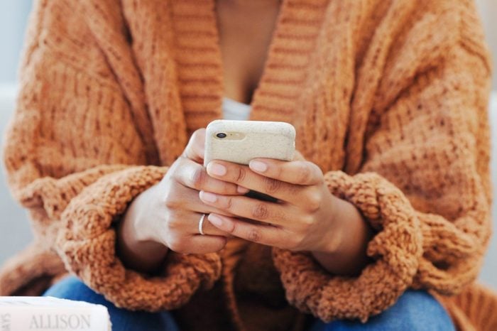 Woman sending a text message, browsing social media or surfing the internet on her phone while sitting in the living room at home. Closeup of the hands of a black female holding wireless technology