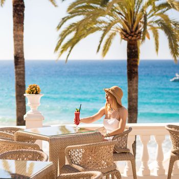 Young Blonde Woman Sitting In The Beach Restaurant Enjoying A Red Cocktail With The Turquoise Sea And Palm Trees And A Yacht In The Background
