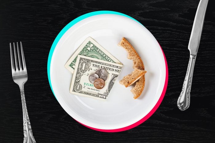 white plate with dollar bill, coins, and crust of bread on black wood background between and fork and knife. the plate has been given blue and pink edges similar to the tiktok logo