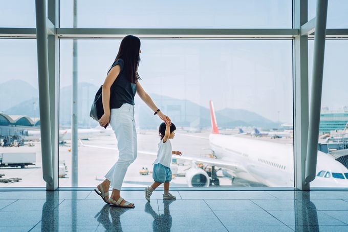 Joyful young Asian mother holding hands of cute little daughter looking at airplane through window at the airport while waiting for departure
