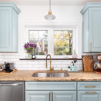A clean and organized light blue kitchen with a granite countertop, gold faucet and light, and a white subway tile backsplash.