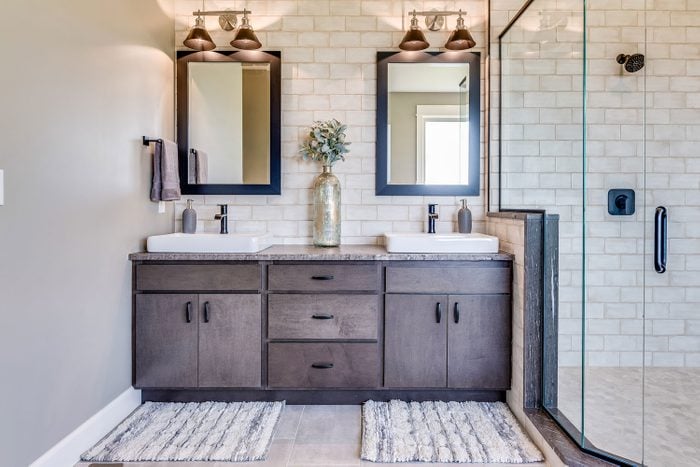Double Vanity With Vessel Sinks against a gray residential bathroom with double mirrors, a walk in shpwer, and bathmats resting in front of each vanity.