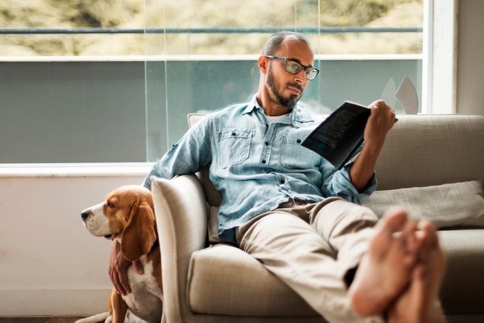 man comfortably sitting on a couch reading a book in one hand and petting his dog with the other hand