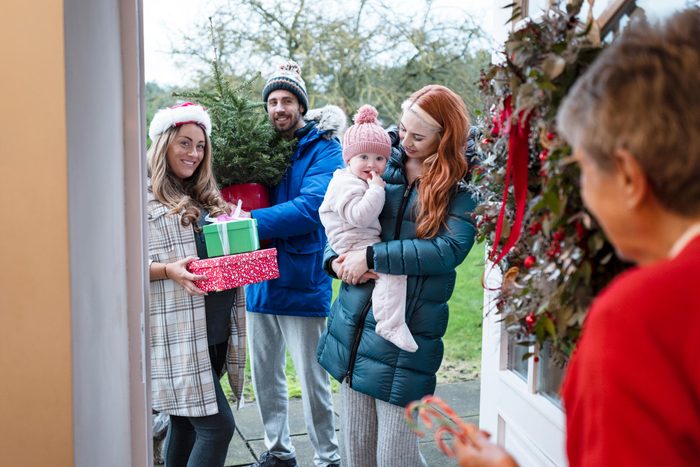 An over the shoulder, medium shot of a senior woman opening her front door to her three grandchildren and her great granddaughter at Christmas. They are standing outside holding gifts and a Christmas tree.