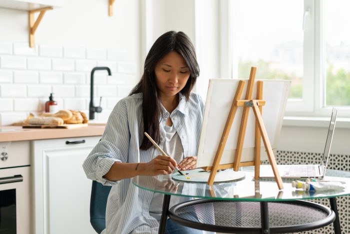 Woman online learning painting skill. Adult asian female oil drawing on easel in kitchen. Following remote art course concept. Improving knowledge at home. Relaxing leisure activity. Artist creative occupation. Mixing colors on palette, copy space