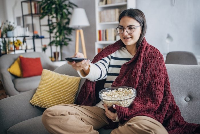 woman sitting on the sofa with a bowl of popcorn pointing a remote at the tv to watch a movie